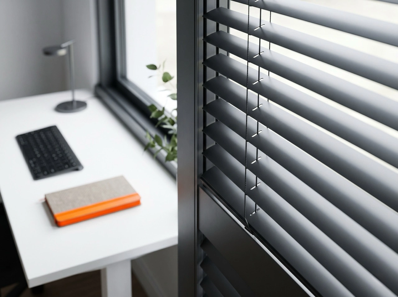 Charcoal aluminium venetian blinds Melbourne in a contemporary office with white desk, black keyboard and orange notebook accent