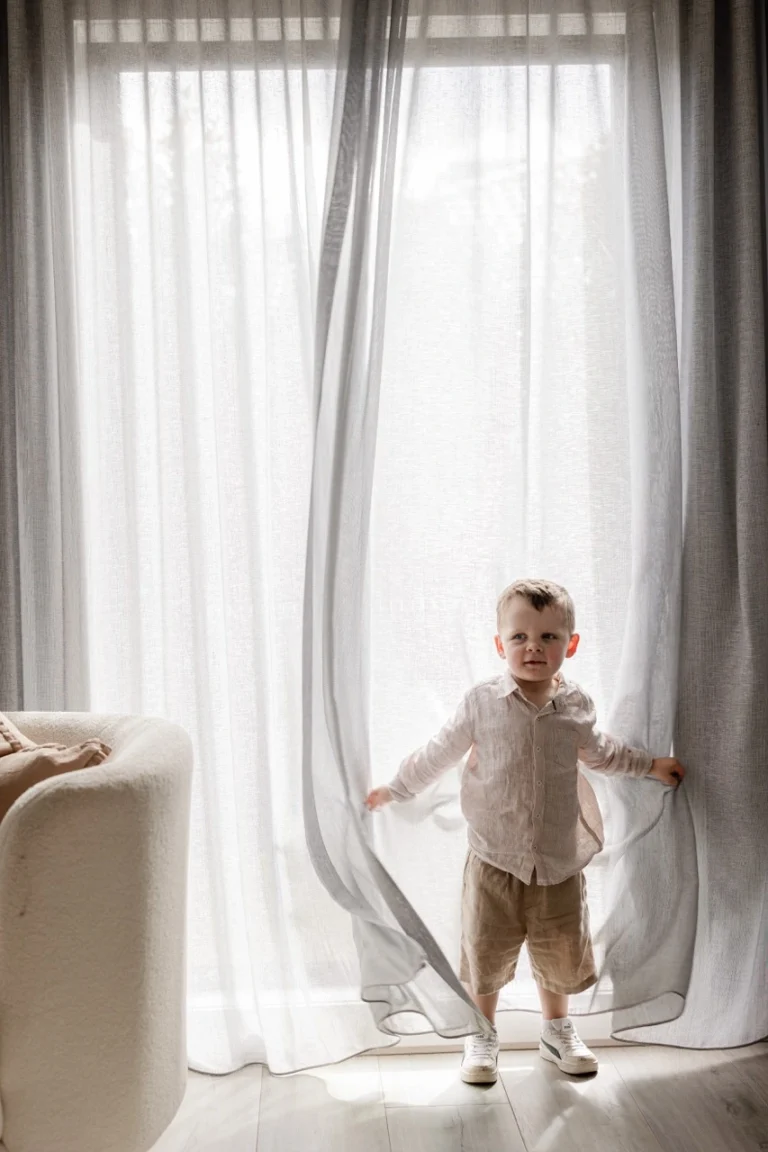 A young boy playfully holding open floor-to-ceiling custom sheer curtains in a sunlit room. The elegant white sheer fabric gently filters bright natural daylight into the home, framed by a light grey side drape.