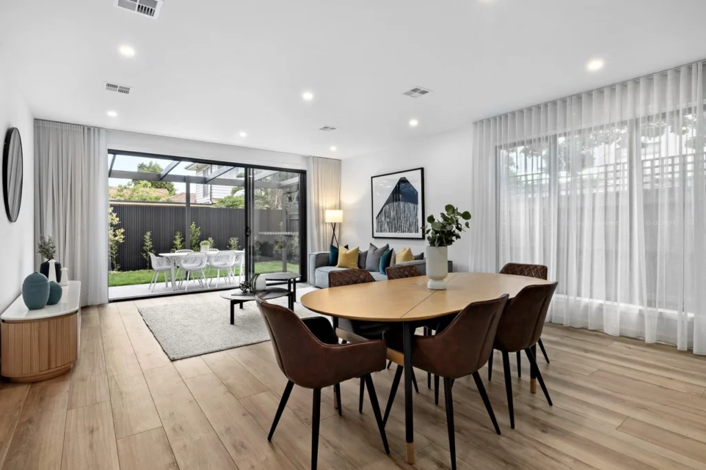 A modern open-plan dining and living room. Large floor-to-ceiling window blinds and glass sliding doors are dressed with custom white sheer curtains, elegantly filtering natural light into the space while maintaining a luxury finish.
