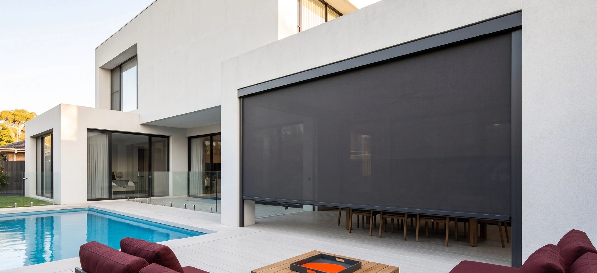 Outdoor blinds Melbourne - extra large charcoal Zipscreen blind covering wide opening on contemporary white two-storey home, swimming pool with blue water, timber deck with deep maroon outdoor lounge seating, orange tray on coffee table, architectural exterior design