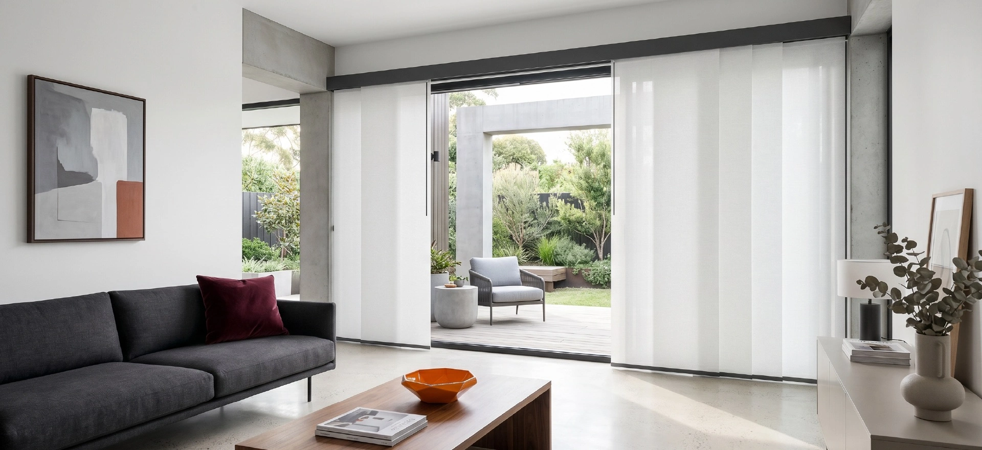 Bright contemporary living room with white sheer panel glide blinds melbourn framing a wide sliding door to a timber deck and garden, styled with a charcoal sofa, abstract artwork, and orange decorative accents.