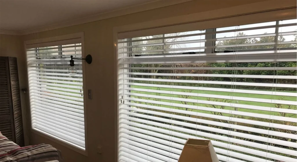 Venetian Blinds Melbourne: The Complete Buying Guide 2 White timber-look Venetian blinds installed across two living room windows overlooking a lawn and garden.